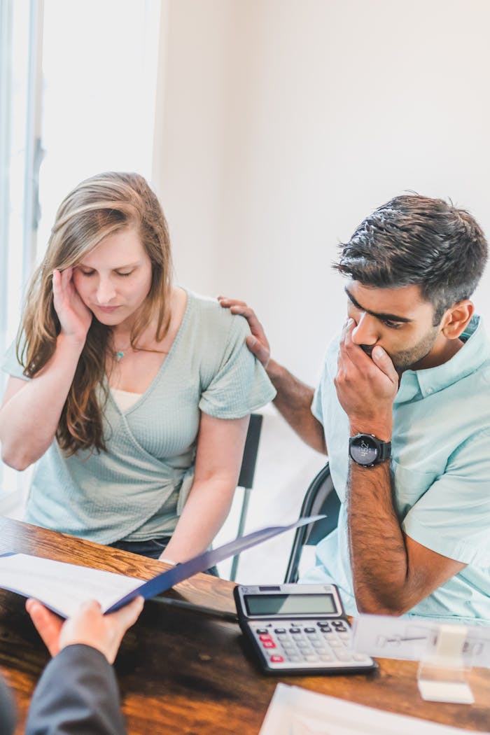 A young couple sits with a real estate agent, reviewing documents and looking concerned about buying a new home.